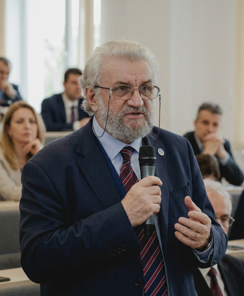 Older man with white hair and beard speaking at a conference holding a microphone